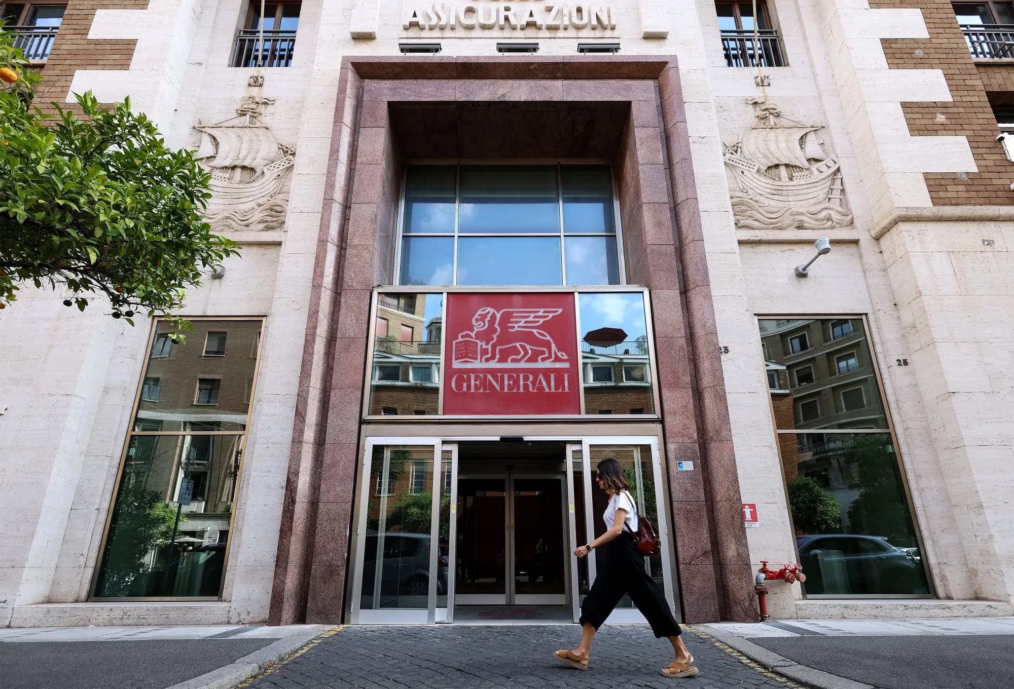 A woman walking past the entrance of the Generali building in Rome, with the red Generali winged lion logo above the doorway