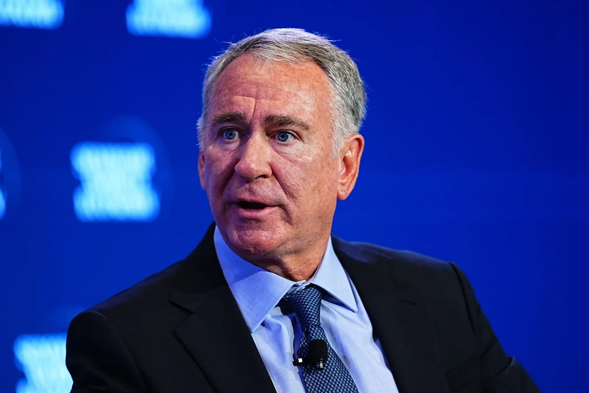 Ken Griffin speaking at a conference, wearing a dark suit and blue tie, with a blue backdrop behind him