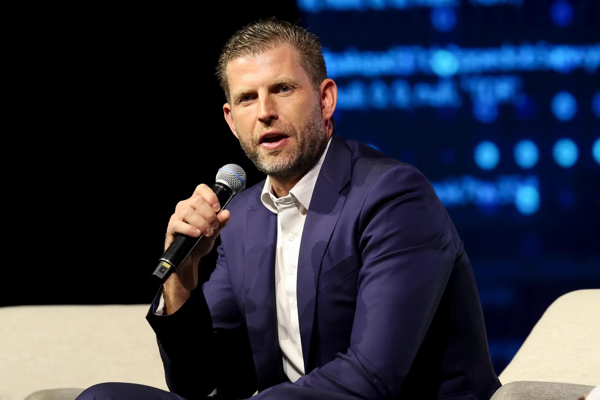 Eric Trump speaking into a microphone while seated on stage, wearing a navy suit, with a blue digital backdrop behind him