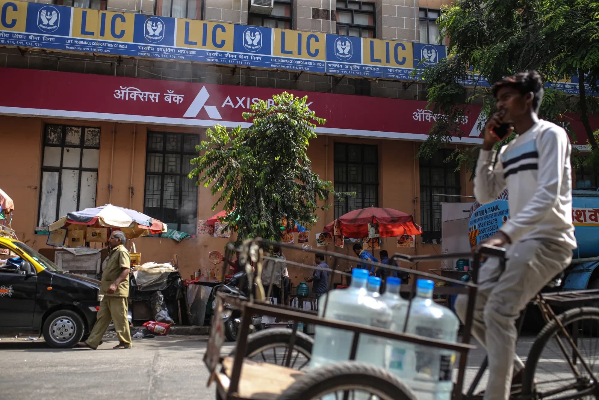 A street scene in India with LIC and Axis Bank signage on a building facade, a man walking past and a water delivery cart in the foreground
