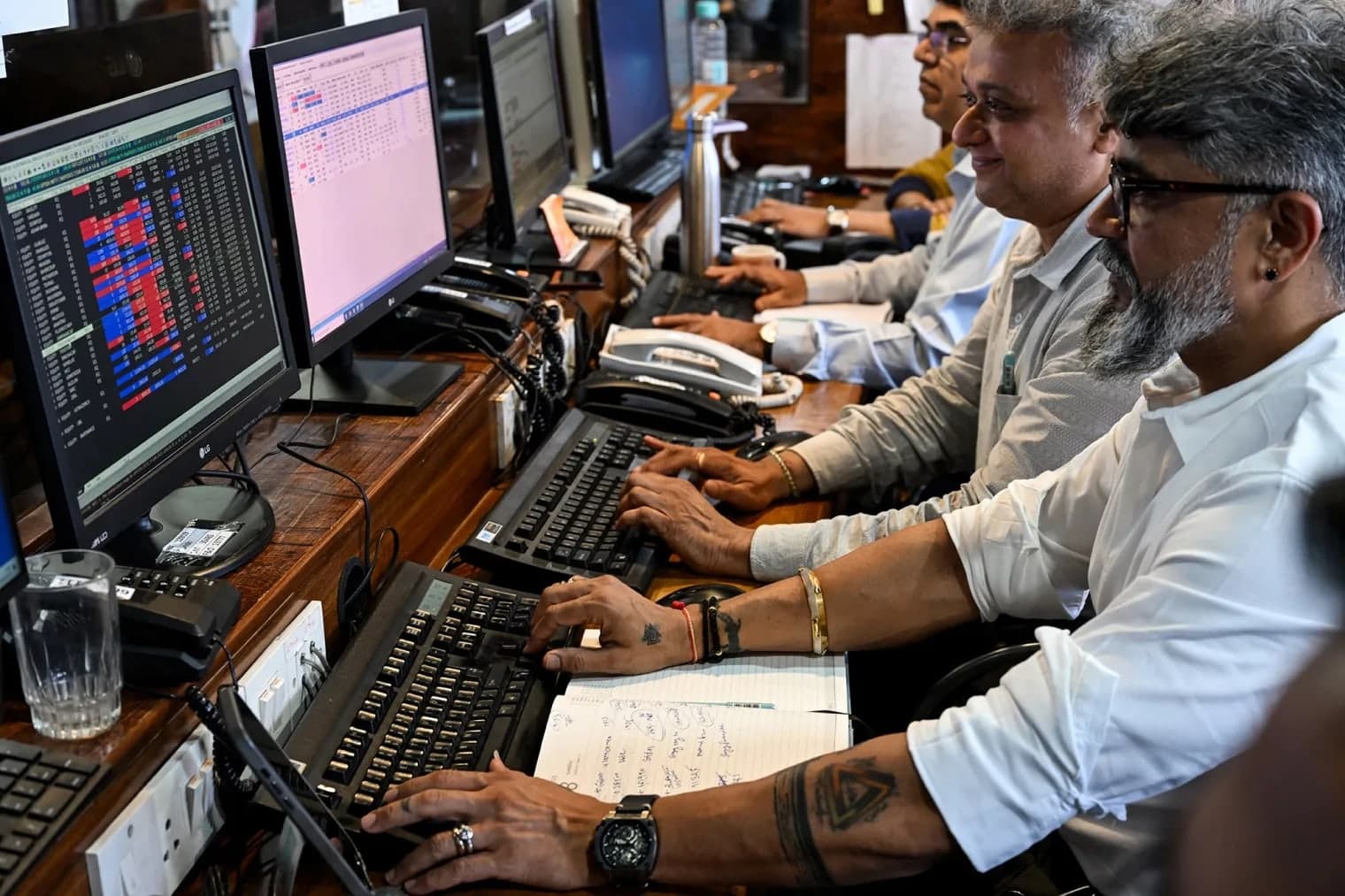 Traders working at a row of desks with multiple monitors showing stock market data and landline phones in an Indian brokerage office