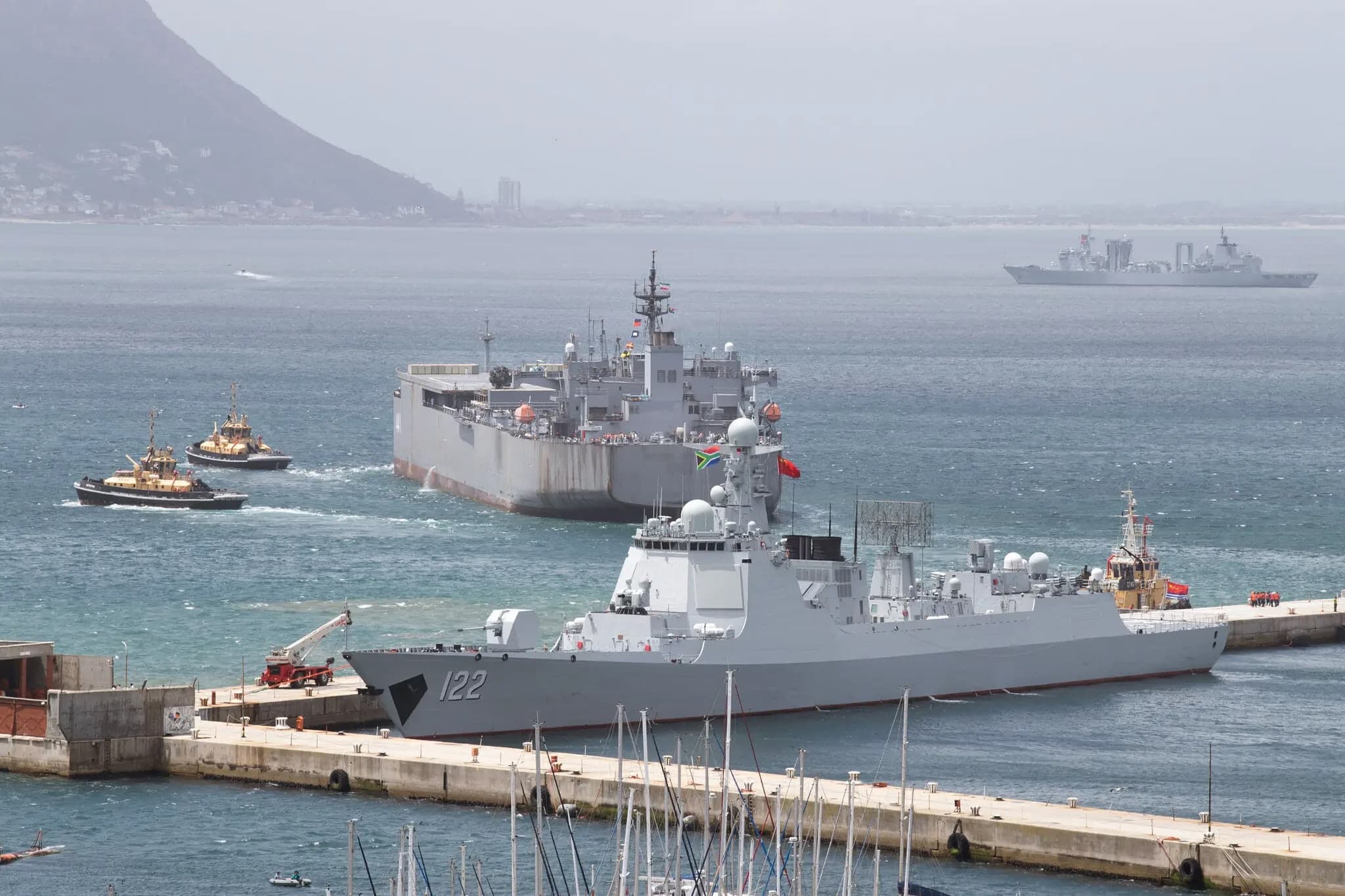 A Chinese navy frigate with hull number 122 docked in Cape Town harbor, with tugboats alongside and a large grey replenishment ship in the background
