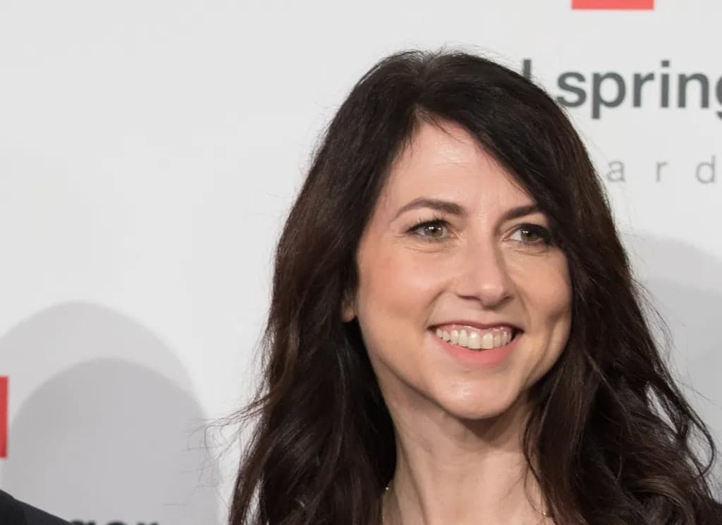 MacKenzie Scott smiling and looking off to the side, with dark wavy hair, photographed at an event in front of a light backdrop