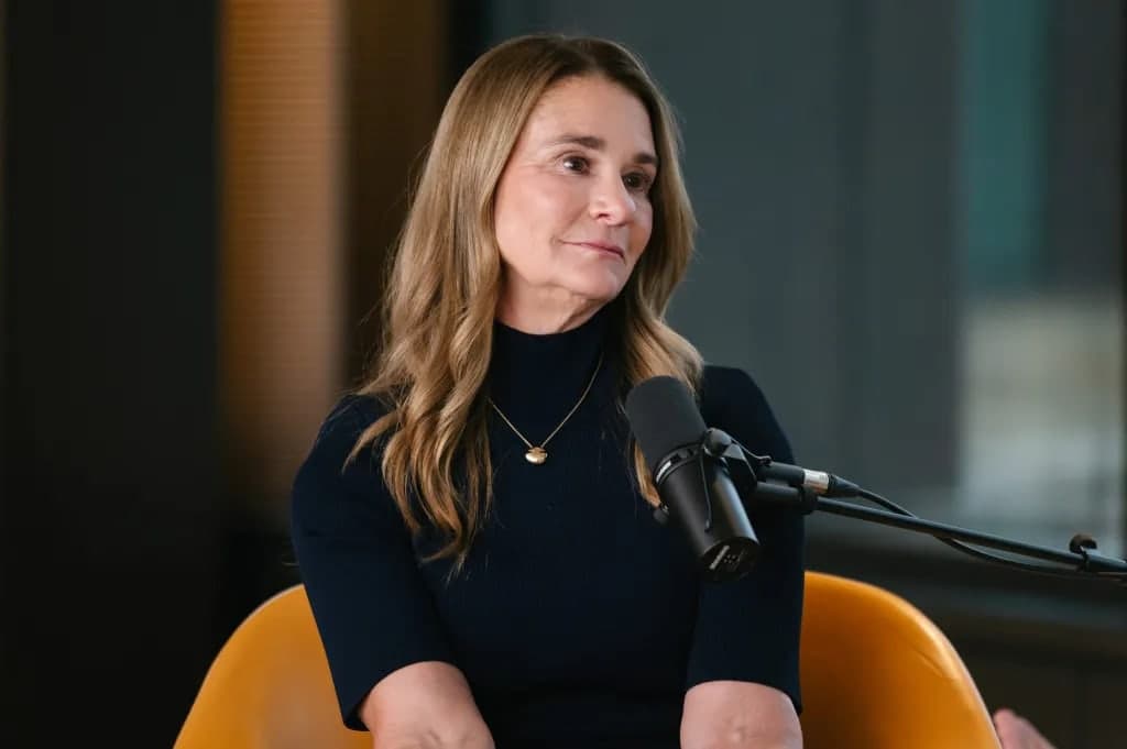 Melinda French Gates seated in an orange chair speaking into a microphone during an interview, wearing a dark sweater and a small gold necklace