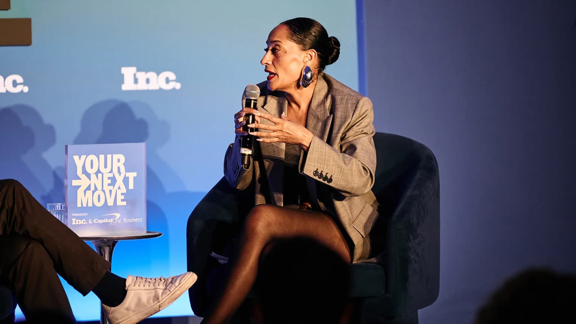 Tracee Ellis Ross speaking into a microphone while seated on stage at an Inc. event, gesturing with her hand, with a sign reading Your Next Move beside her